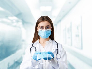 Yüz maskeli bir kadın doktor. Beyaz üniformalı orta yaşlı kadın doktor mavi bulanık koridor hastane geçmişine yüz maskesi takıyor..