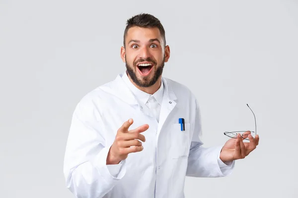Healthcare workers, coronavirus, covid-19 pandemic and insurance concept. Enthusiastic handsome doctor in white coat, pointing finger at camera and smiling amused, surprised, excited hear good news — Stock Photo, Image