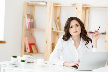 Modern bir ofis masasında dizüstü bilgisayarla çalışan ve bir şeyler düşünen çekici genç bir kadın..