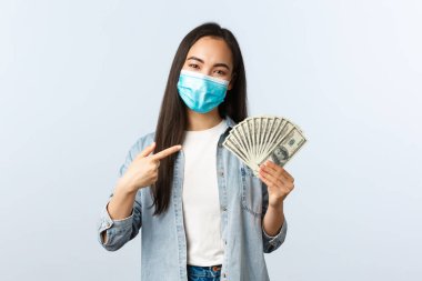 Sosyal mesafeli yaşam tarzı, covid-19 pandemik iş ve heyecan konsepti. Genç mutlu Asyalı serbest çalışan kadın Coronavirus üzerinde çalışıyor, paralı dolarları işaret ediyor.