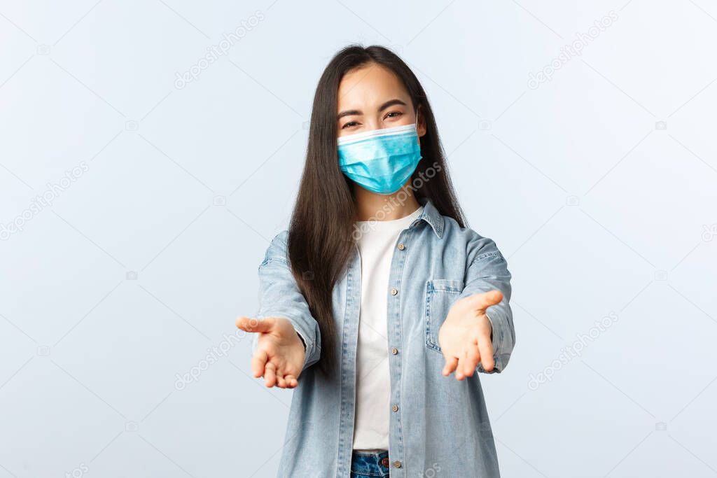 Social distancing lifestyle, covid-19 pandemic everyday life and leisure concept. Friendly lovely asian woman in medical mask reaching hands forward to take something or hold, white background