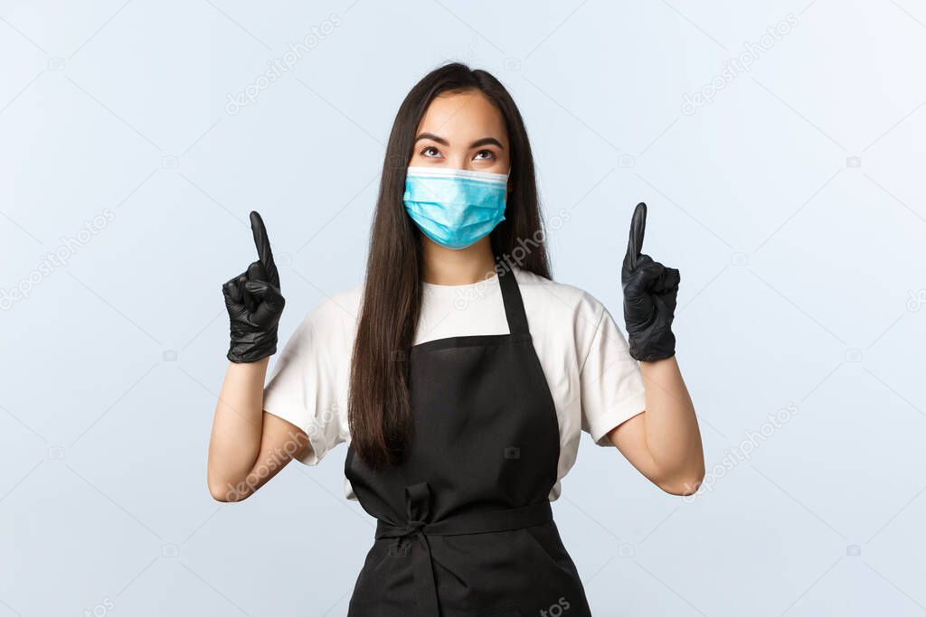 Covid-19 pandemic, social distancing, small business, preventing virus concept. Happy smiling asian coffee shop owner, barista in medical mask and gloves pointing looking at upper banner, top promo