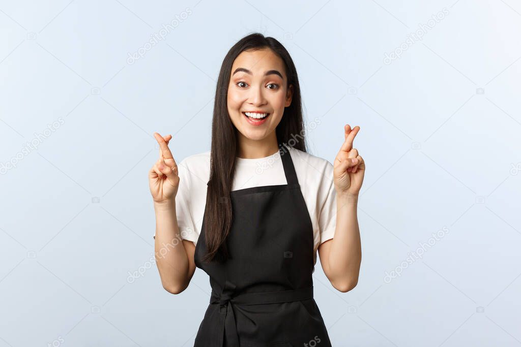 Coffee shop, small business and startup concept. Optimistic cute asian waitress cross fingers good luck. Upbeat barista in black apron making wish, praying or pleading