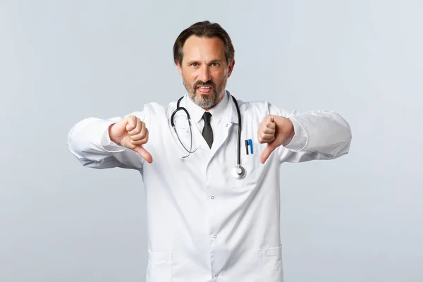 Covid-19, ξέσπασμα του ιού της στέψης, επαγγελματίες υγείας και πανδημία. Θυμωμένος απογοητευμένος άντρας γιατρός με λευκό παλτό, παραπονιέται για έντονη απέχθεια, δείχνει τους αντίχειρες προς τα κάτω — Φωτογραφία Αρχείου
