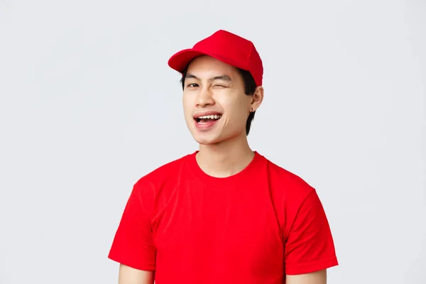 Παράδοση, ανέπαφες παραγγελίες και concept αγορών. Enthusiastic ασιατική courier σε κόκκινο στολή t-shirt και καπέλο, χαμογελώντας διασκεδάζει και κλείσιμο του ματιού στον πελάτη, συνιστούμε την υπηρεσία, στέκεται γκρι φόντο — Φωτογραφία Αρχείου