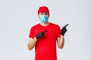 Covid-19, kendini karantina, online alışveriş ve nakliye konsepti. Üzgün ve canı sıkkın teslimatçı, rahatsız yüzüyle bakıyor ve işaret ediyor. Üniforma giyiyor, tıbbi maske ve eldiven takıyor.