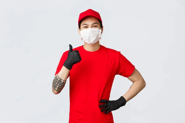 Dont worry your parcel in safe in our hands. Confident smiling asian delivery guy in red uniform, protective medical mask and gloves, thumbs-up in approval, say yes, assure all good, no problem — Stock Photo, Image