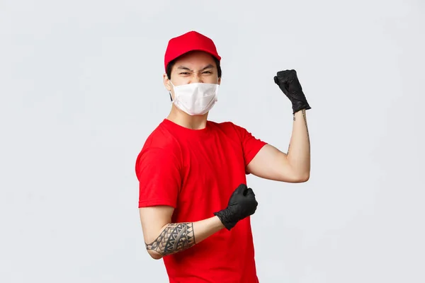 Nadšený asijský doručovatel v červené uniformě s čepicí a tričkem, s ochrannými rukavicemi a lékařskou maskou, posilující sebedůvěrou, pěst pumpa, oslavující úspěch, připraven k předání balíků klientům — Stock fotografie