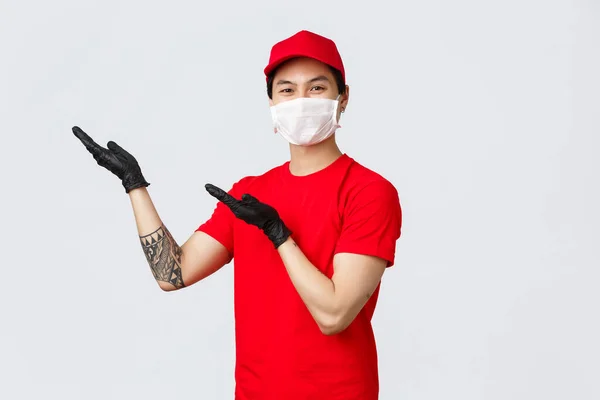 Hier sind Sie. Freundlicher asiatischer Zusteller in roter Mütze, T-Shirt, Trägerdienst vorstellen, Bestellverfolgungsanwendung, Zeigefinger oben links. Kurier in medizinischer Maske und Schutzhandschuhen — Stockfoto