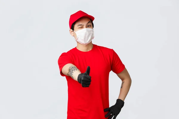 Alegre mensajero asiático en gorra roja y camiseta, usar máscara médica y guantes de protección, guiño y pulgar hacia arriba en la aprobación, recomiendan un servicio de entrega de buena calidad. Empleado promover la empresa logística rápida —  Fotos de Stock