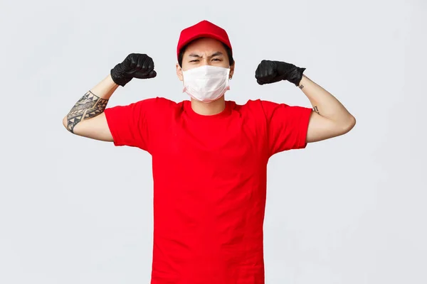 Forte asiatique livreur gars en bonnet rouge, t-shirt montrer sa force, flexion biceps vantardise muscles comme transporter de grandes boîtes au travail. Courrier livrer facilement des colis lourds. Concept de transporteurs et d'achats — Photo
