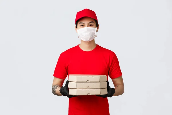 Food delivery, contactless shopping online concept. Friendly asian delivery guy in red uniform in medical mask and gloves standing clients doorstep, deliver pizzas, courier