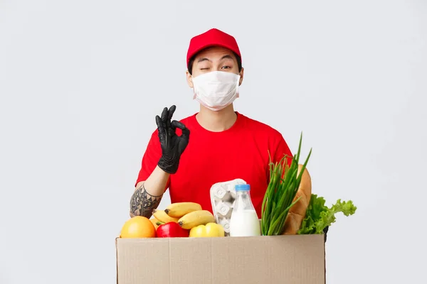 Online shopping, food delivery and coronavirus pandemic concept. Smiling asian courier in uniform show okay sign, give client his order, fresh groceries from online shop, wear medical mask