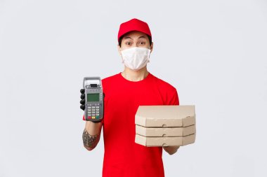 Temassız teslimat, güvenli alım, koronavirüs konsepti sırasında alışveriş. Tıbbi maske ve eldiven takan hevesli pizzacı. Müşterinin kredi kartıyla ödeme yapmasını sağlamak için POS terminalini kullan.