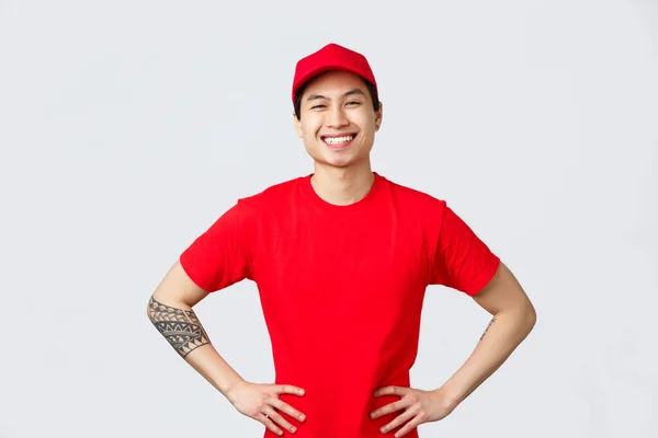 Expresní dodávka, expedice a logistika koncepce. Usmívající se charismatický asijský muž donáška muž v červené čepici, tričko, držet ruce v bok, vypadající potěšený, připraven pomoci klientovi, rozdávat zákaznické balíčky — Stock fotografie