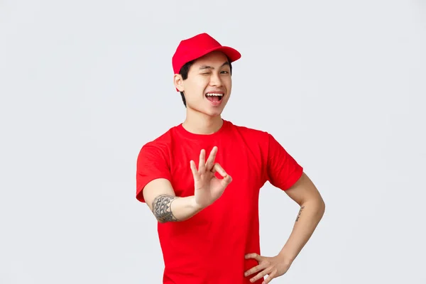 A legjobb üzlet a piacon. Jóképű mosolygós ázsiai futár piros sapkában és pólóban biztosítja a legjobb minőségű árut, a csomagok biztonságát. Futár show rendben, jóváhagyási jel, kacsintós pimasz kamera — Stock Fotó