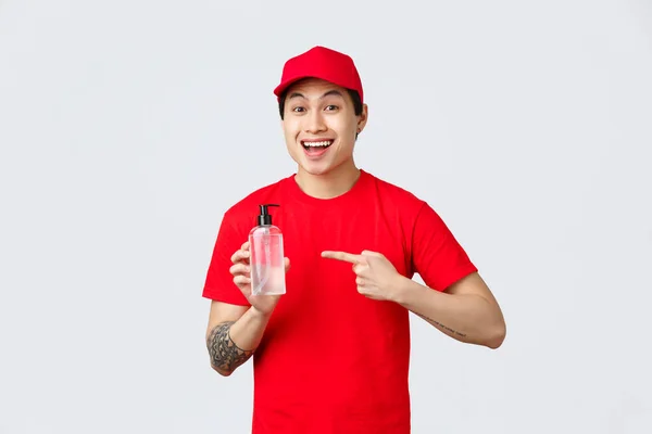 Karanténa, bezpečný bezkontaktní doručovací a nákupní koncept. Veselý usměvavý asijský kurýr v červené čepici a tričku, ukazující na dezinfekci rukou, podporující bezpečné nakupování, význam prevence šíření viru — Stock fotografie