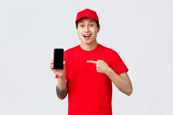 Dodací aplikace, on-line nákupní a přepravní koncept. Překvapený asijský kurýr v červené čepici a tričku, ukazující prstem na aplikaci mobilního telefonu, zobrazující obrazovku smartphonu s pobavenou tváří — Stock fotografie