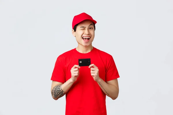 Amigável sorrindo asiand elivery homem em boné vermelho e uniforme t-shirt, piscar alegre, recomendo o uso de cartão de crédito, comprar contactless durante o surto de vírus. Correio dar ordem de aconselhamento on-line sobre coronavírus — Fotografia de Stock