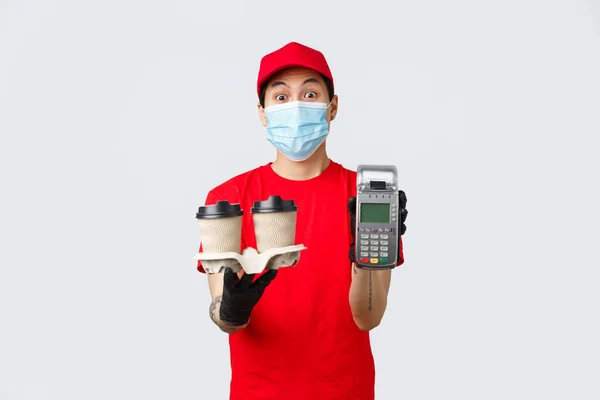 Contactless delivery, safe purchase and shopping during coronavirus concept. Asian courier or fast food employee showing POS terminal and coffee, paying terminal to customer, grey background