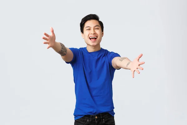 Veselý přátelsky vypadající mladý muž student, asijský muž v modrém tričku s tetováním, natahující ruce k fotoaparátu pro objetí, chtějí objetí nebo objetí, pozdrav drahý přítel, šedé pozadí — Stock fotografie