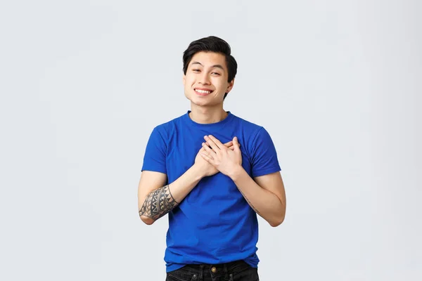 Доторканий і вдячний красивий азіатський чоловік у синій футболці з татуюваннями, зворушливим серцем і посмішкою, цінуючи допомогу, будучи задоволеним або в захваті, отримує сердечний комплімент, сірий фон — стокове фото