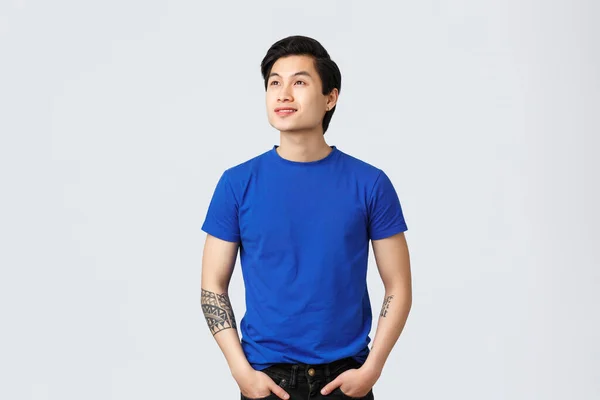 Sonhador, pensativo bonito asiático queer guy em azul t-shirt, olhando para a frente algo, olhando para o canto superior esquerdo com deleitado, sorridente rosto, lendo banner ou promo, fundo cinza — Fotografia de Stock