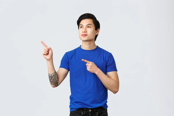 Dromerige jonge mannelijke Aziatische student, man met ambities kijken en wijzen linksboven hoek met enthousiaste, tevreden uitdrukking. Taiwanese man in blauw t-shirt staande grijze achtergrond — Stockfoto