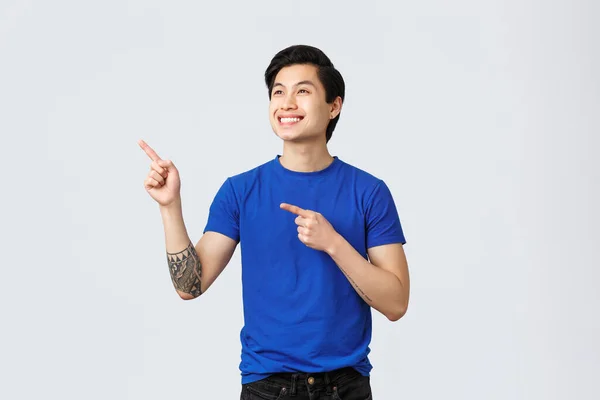 Carefree smiling asian guy in blue t-shirt, gazing at upper left corner, pointing fingers at promo or banner. Man on self-quarantine during covid-19 look out window, standing grey background — Stock Photo, Image