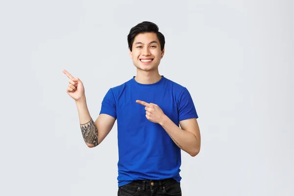 Bonito jovem asiático homem em azul t-shirt, tatuagem no braço, apontando dedos canto superior esquerdo, sorrindo como introduzir produto de propaganda, de pé fundo cinza alegre — Fotografia de Stock