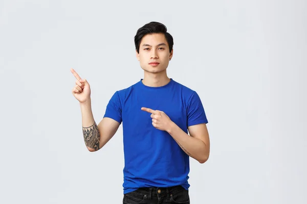 Různé emoce, životní styl lidí a reklamní koncept. Odhodlaný seriózní asijský muž v modrém tričku, ukazující prsty v levém horním rohu, ukazující prapor, doporučující produkt nebo službu — Stock fotografie