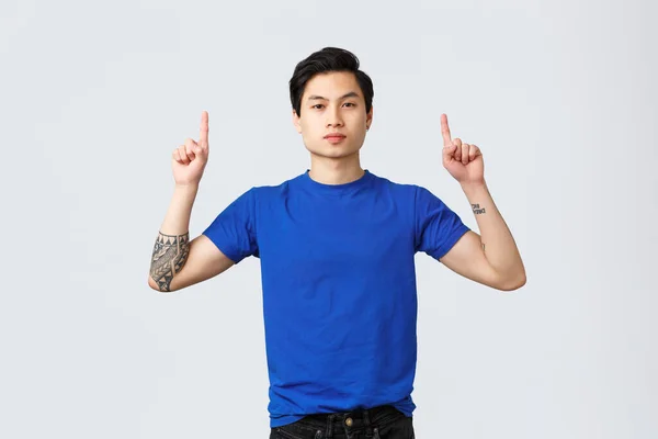 Différentes émotions, style de vie des gens et concept de publicité. Homme asiatique confiant et sérieux en t-shirt bleu avec tatouages, pointant du doigt vers le haut, informant les clients, montrant la voie à la bannière, fond gris — Photo