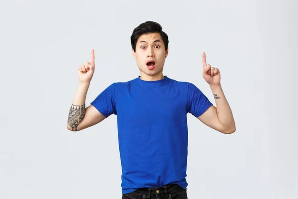 Different emotions, people lifestyle and advertising concept. Surprised and astonished asian male student, guy in blue t-shirt gasping amazed, pointing fingers up at banner, grey background — Stock Photo, Image