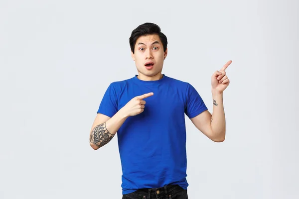 Verschillende emoties, mensen levensstijl en reclame concept. Geschokt en zenuwachtig jonge aziatische man in blauw t-shirt wijzende vingers rechter bovenhoek. Gasping guy tonen spannend nieuws — Stockfoto