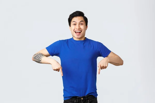 Különböző érzelmek, emberek életmód és reklám koncepció. Lelkes, jóképű ázsiai férfi kék pólóban, ujjal mutogatva lefelé és mosolyogva, ahogy bejelentik a jó hírt, szürke háttér — Stock Fotó