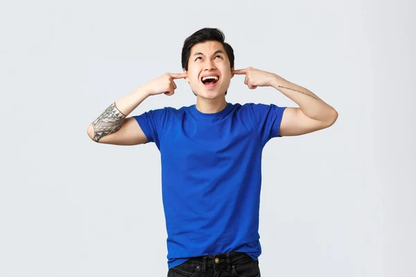People different emotions, lifestyle and casual concept. Angry and pissed-off asian man in blue t-shirt, shut ears and complaining loud neighbours upstairs, staring furious up, stand grey background — Stockfoto