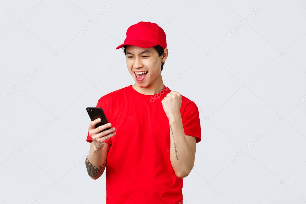 Happy young asian delivery guy in red uniform t-shirt and cap, reading good news smartphone screen, make fist pump chanting great bonuses or success. Courier say yes, tracking orders
