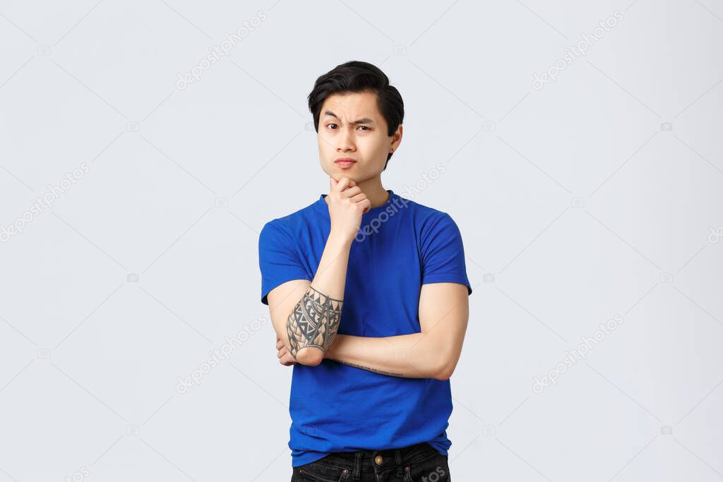 Skeptical, serious-looking asian man in blue t-shirt with tattoos, listening suspicious to person, frowning, touch chin thoughtful, thinking, making decision, express disbelief, grey background