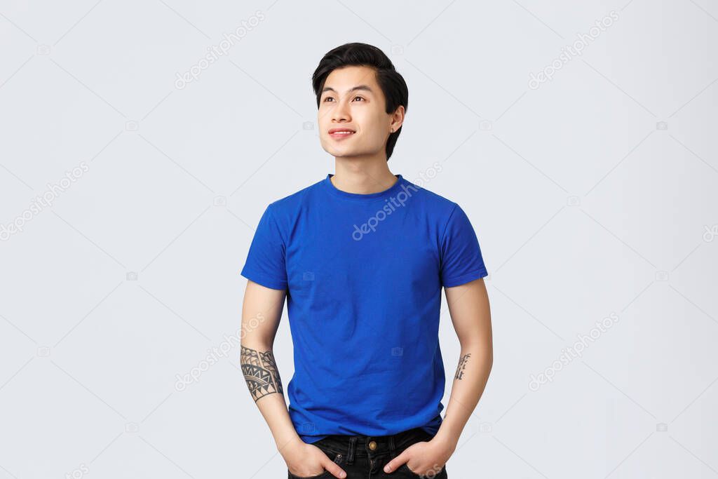 Dreamy, thoughtful handsome asian queer guy in blue t-shirt, looking forward something, gazing at upper left corner with delighted, smiling face, reading banner or promo, grey background