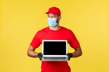 Covid-19, güvenli alışveriş, hızlı teslimat ve sipariş izleme konsepti. Kırmızı üniformalı yakışıklı kurye, tıbbi maske ve eldivenler, göz kırpan kamera ve dizüstü bilgisayar ekranında sayfa sitesi