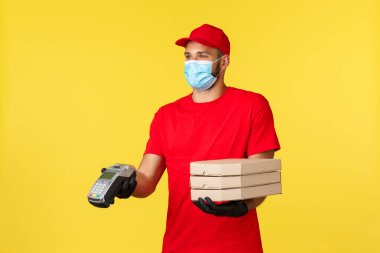 Yemek teslimatı, izleme emirleri, covid-19 ve kendini karantina konsepti. Tıbbi maskeli ve kırmızı üniformalı güler yüzlü kurye müşterilere ödeme terminali ve pizza veriyor.