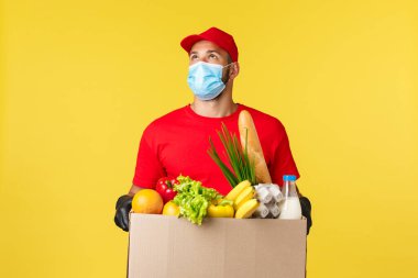 Paket servis, yiyecek ve içecekler, covid-19 karantina ve yiyecek konsepti. Kurye binaya bakıyor. Bakkal kutusunu eve ya da ofise getiriyor. Koruyucu tıbbi maske ve eldiven takıyor.
