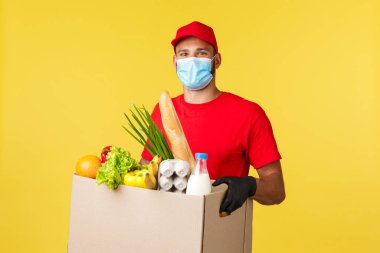 Paket teslimat, COVid-19 karantina ve market konsepti. Tıbbi maske takan neşeli kurye ve kırmızı üniformalı. Müşteri evine yiyecek siparişi verin.