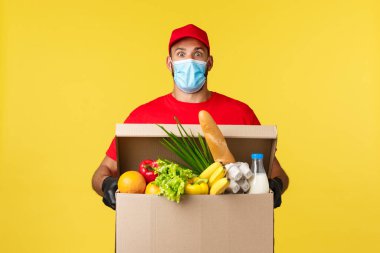 Paket teslimat, COVid-19 karantina ve market konsepti. Şaşkın kurye tıbbi maske, üniforma kırmızı başlık ve tişört, şaşkın şaşkın bakışlar, eve yemek siparişi kutusu getirmek, sarı arka plan