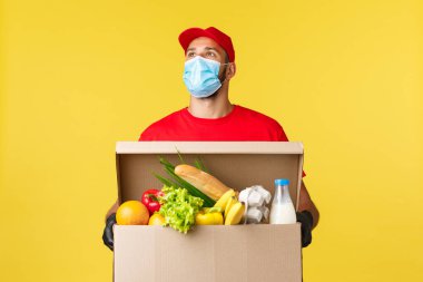 Paket teslimat, COVid-19 karantina ve market konsepti. Kırmızı şapkalı ve t-shirt 'lü genç kurye, tıbbi maske, düşünceli, müşteri dairesini arayan, içinde yemek siparişi olan bir kutu.