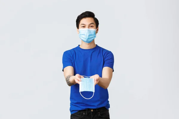 Different emotions, social distancing, self-quarantine on coronavirus and lifestyle concept. Handsome asian male volunteer, guy in personal protective equipment give you medical mask during covid-19