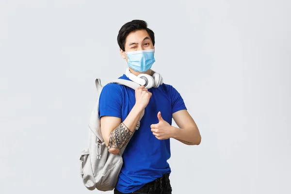 Tillbaka till skolan, studier på covid-19, utbildning och universitetsliv koncept. Snygg hipster kille, asiatisk student i medicinsk mask, visa tummen upp som huvudet klasser med ryggsäck och hörlurar — Stockfoto