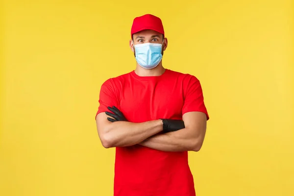 Consegna espressa durante pandemia, covid-19, spedizione sicura, concetto di shopping online. Sorpreso corriere maschio in cappuccio uniforme rossa e t-shirt, petto braccia incrociate, maschera medica, stand sfondo giallo — Foto Stock