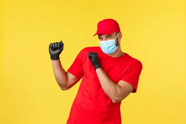 Entrega expressa durante pandemia, covid-19, transporte seguro, conceito de compras on-line. Correio sorridente despreocupado em uniforme vermelho, máscara médica e luvas, dança, promo da empresa de logística — Fotografia de Stock