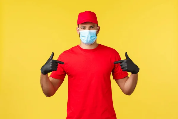 Express levering tijdens pandemie, covid-19, veilige verzending, online shopping concept. Jonge koerier in rood uniform, medisch masker en handschoenen op zichzelf gericht, zorgen voor snelle levering van pakketten — Stockfoto